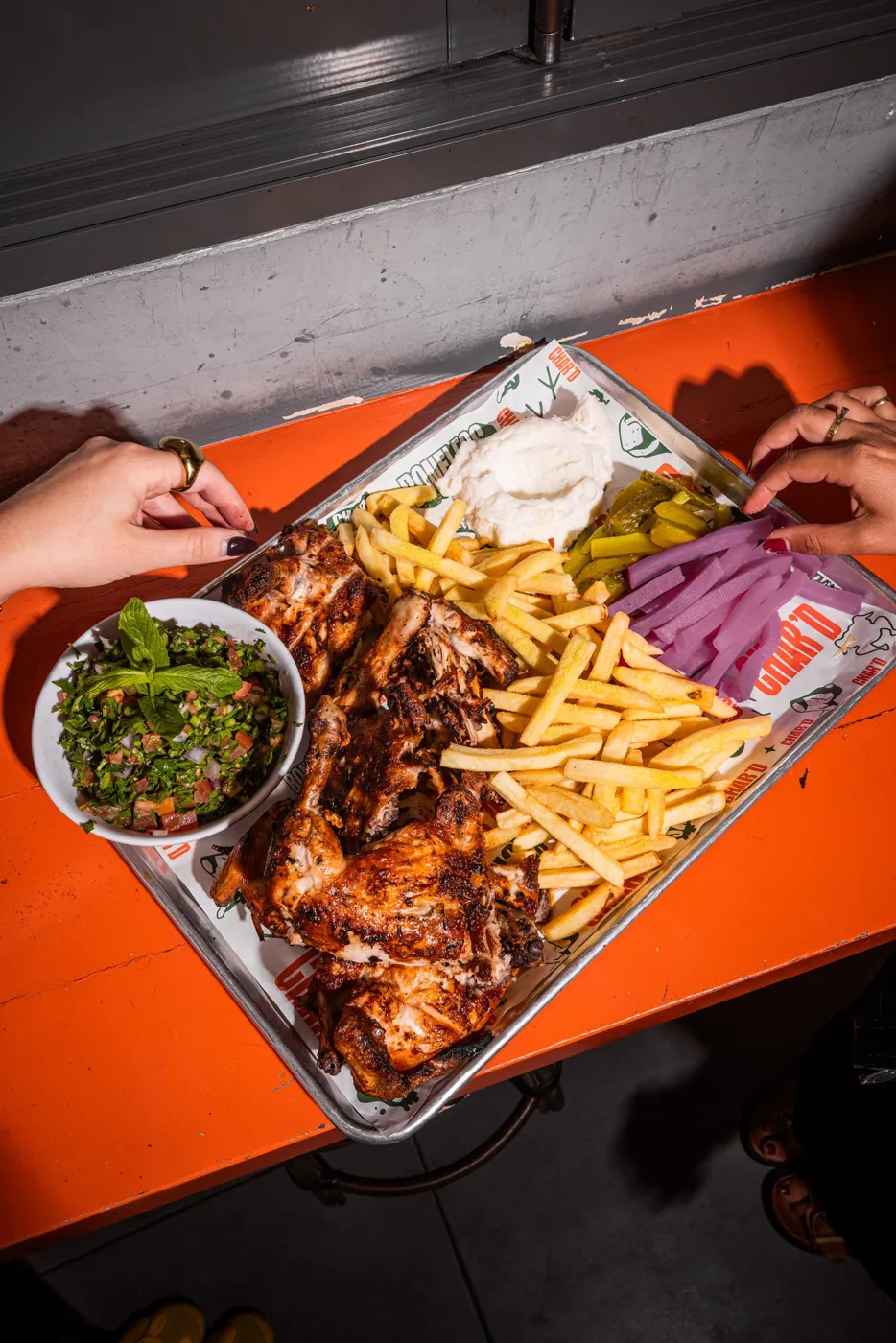 Hands reaching into a Char’d platter of charcoal chicken, chips, salads and garlic sauce